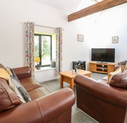 A living room with brown leather sofas a wooden coffee table a television on a stand and a window with floral curtains at The Long Barn in Morton on the Hill near Lenwade