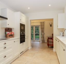 A kitchen with white cabinets an oven a wicker basket and a sink with a window showing a plaid armchair and glass doors at Lavender Cottage in Market Weighton