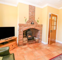 A room with a brick fireplace wood stove television on a wooden stand armchair and partially open wooden door at The Meadows Cottage in Billingford near Bawdeswell