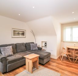 A sitting area with a gray sofa and two pillows a wooden coffee table a small wooden dining table with two chairs and a window with blinds at Shephard's Retreat in Charlton Mackrell near Somerton