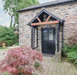 A stone building entrance with a black door and wooden canopy surrounded by plants at The Old Barn in Llanferres near Ruthin