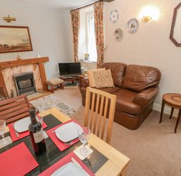 A living room with a dining table set for four a brown leather sofa a fireplace and a small television at Blacksmith Cottage in Appleby-in-Westmorland