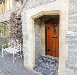 A wooden door with a heart decoration in a stone wall next to a white metal bench on a paved area at Bears Cottage in Bretforton near Broadway