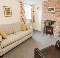 A living room with a beige sofa and cushions a brown armchair a brick fireplace with wood stove a window with floral curtains and framed pictures at Bears Cottage in Bretforton near Broadway