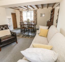 A living room with a beige sofa and brown armchair next to a wooden dining table with chairs and floral curtains at Bears Cottage in Bretforton near Broadway