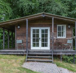 A wooden cabin with a porch two chairs a small table and steps leading to a gravel path at Lakeside Cabin in Sheldon Blackdown Hills