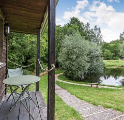 A wooden porch with a green table and chair overlooking a pond and trees at Lakeside Cabin in Sheldon Blackdown Hills