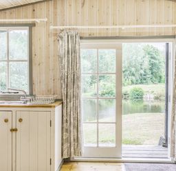 A kitchen area with a sink and dish rack next to a glass door overlooking a pond and greenery at Lakeside Cabin in Sheldon Blackdown Hills