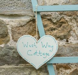 A heart shaped sign reading West Wing Cottage hanging on a blue wooden frame against a stone wall at West Wing Cottage in Hexham