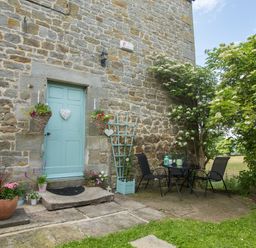 An outdoor patio area with a blue door on a stone wall two black chairs and a table with plants and flowers at West Wing Cottage in Hexham