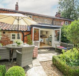 A garden patio with a table and chairs under an umbrella beside an open door to a kitchen at Bridge Cottage in Aylsham