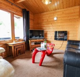 A living room with wooden walls and ceiling a black leather sofa a cream armchair a red glass coffee table a television and nested wooden tables at Heathcliff in Northallerton