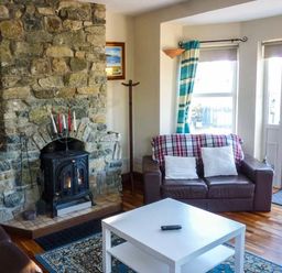 A living room with a stone fireplace a brown leather sofa a white coffee table and windows with blue curtains at Ti Sheamuis in Cashel near Carna County Galway