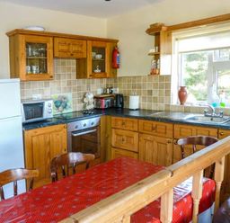 A kitchen with wooden cabinets a white refrigerator a microwave stove and a red tablecloth on the dining table at Ti Sheamuis in Cashel near Carna County Galway