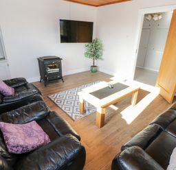 A living room with black leather sofas and purple pillows a wooden coffee table a patterned rug a wall-mounted tv a stove and a potted plant at Aras Iosgan in Carraroe County Galway