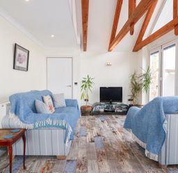 A living room with two sofas covered with blue throws a small wooden table a television on a stand with books wooden ceiling beams and large windows at Chapel Green Studio 2 in Polgooth
