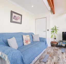 A living room with a blue covered sofa three cushions a potted plant a TV on a glass stand and a wall painting at Chapel Green Studio 2 in Polgooth