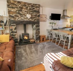 A living room with a stone fireplace a wood-burning stove brown sofas a dining table with chairs and a kitchen area at Snowdon View in Llanberis