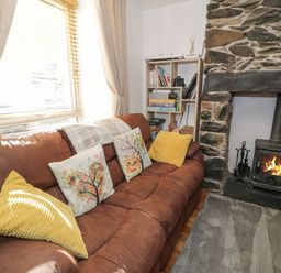 A living room with a brown sofa with decorative cushions next to a stone fireplace with a wood stove at Snowdon View in Llanberis