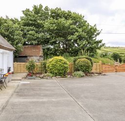 A concrete driveway with outdoor metal table and chairs next to a thatched white building and wooden gates in a rural landscape at Whispering Willows - The Thatch in Carndonagh County Donegal
