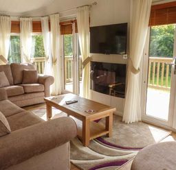 A living room with brown sofas a wooden coffee table a wall mounted TV and large windows with curtains at Morgan Lodge in Hewish near Weston-super-Mare