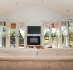 A living room with beige sofas a coffee table a wall mounted television and fireplace with large windows and sheer curtains at Harvester Lodge in Hewish near Weston-super-Mare