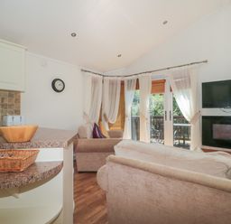 A living room with two sofas a wall-mounted television and fireplace and a kitchen counter with a wooden bowl at Harvester Lodge in Hewish near Weston-super-Mare