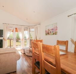 A dining area with a wooden table and chairs next to a living room with a beige sofa and a window with sheer curtains at Harvester Lodge in Hewish near Weston-super-Mare