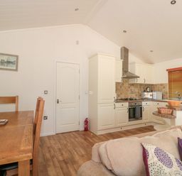 An open plan kitchen and dining area with wooden dining table and chairs beige kitchen cabinets and a window with blinds at Harvester Lodge in Hewish near Weston-super-Mare
