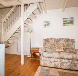A living area with a floral sofa a wooden side table a floor lamp a staircase and framed pictures on the wall at The Ark Cottage in St Blazey