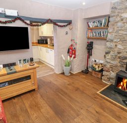 A living room with a wood stove fire a wooden cabinet with framed photos a television and an open doorway to a kitchen at Stable Cottage in Pembroke