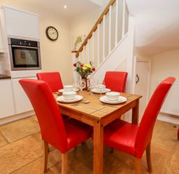 A dining area with a wooden table set for four with red chairs and a kitchen with white cabinets and an oven at Cow Byre Great Tew near Chipping Norton