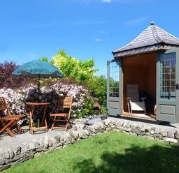 A garden with a stone patio table and chairs with an umbrella and a small wooden gazebo with open doors at High House Newton in Cartmel near Cartmel