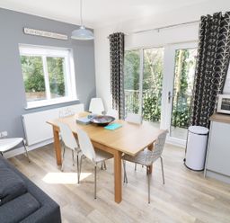 A dining area with a wooden table and six white chairs next to a gray sofa and glass doors at Wysteria Cottage in Windermere