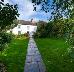 A stone pathway leading to a white cottage with green doors and chimney surrounded by grass and trees at Fern Cottage in Aberporth