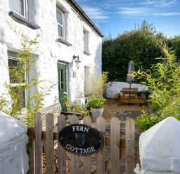 An outdoor area with a wooden gate labeled Fern Cottage white cottage walls green door wooden bench potted plants and a wooden picnic table with a closed umbrella in Aberporth