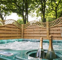 A bubbling outdoor hot tub with two bottles of champagne and two empty glasses in a wooden fenced area with trees at Pond Farm Woodlands in Ugthorpe