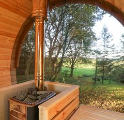 The interior of a wooden sauna with a stove and rocks and a large window showing trees and grass outside at Pond Farm Woodlands in Ugthorpe