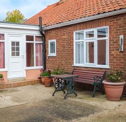 An outdoor patio area with a wooden bench and metal table next to a brick house with white windows and flower pots at Glenhaven in Sweffling near Snape