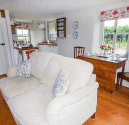 A living area with a cream sofa, wooden floor, wooden table with chairs, and kitchen in the background at Rose Cottage in Welsh Newton