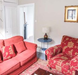 A living room with a red sofa and patterned armchair a round side table with a lamp and a framed picture on the wall at Woodland Cottage in Malvern