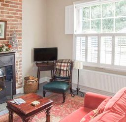 A living room with a fireplace a red sofa a wooden coffee table a chair with a blanket a television and a window with shutters at Woodland Cottage in Malvern
