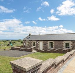 A brick house with a paved walkway and fenced grass area with playground equipment at Home Stead Cottage in Embleton