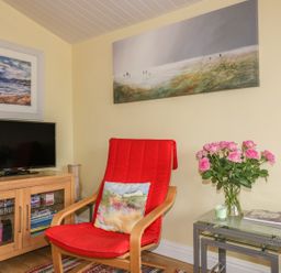 A sitting area with a red cushioned chair a wooden TV stand with a television and a side table with pink flowers at Tiggy Winkle in St Merryn near Padstow