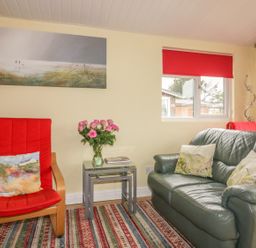 A living room with a red armchair and a leather sofa with cushions and a side table with flowers at Tiggy Winkle in St Merryn near Padstow