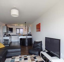 An open living room and kitchen with a black sectional sofa with yellow pillows a patterned rug a flat screen tv on a white stand and a round black dining table at Tehidy in Charlestown