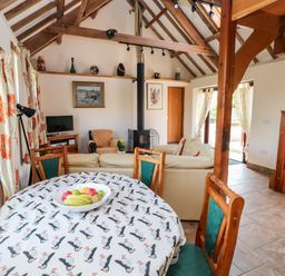 A living room with a dining table covered with a patterned tablecloth a bowl of fruit sofas a wood burning stove wooden staircase and patterned curtains at The Long Barn in Hasguard near Little Haven
