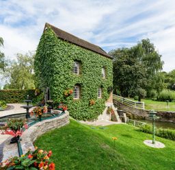 An ivy-covered stone building next to a circular fountain and a grassy area with trees and a small canal at Grafton Mill Grafton Flyford near Pershore
