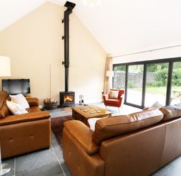 A living room with brown leather sofas a wood stove and large windows at Hen Gelligemlyn - The Annexe in Ganllwyd