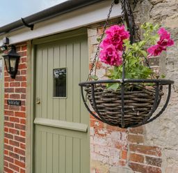 A hanging basket with pink flowers outside a green door on a brick and stone wall at The Old Cart Shed in Lyneham near Royal Wootton Bassett
